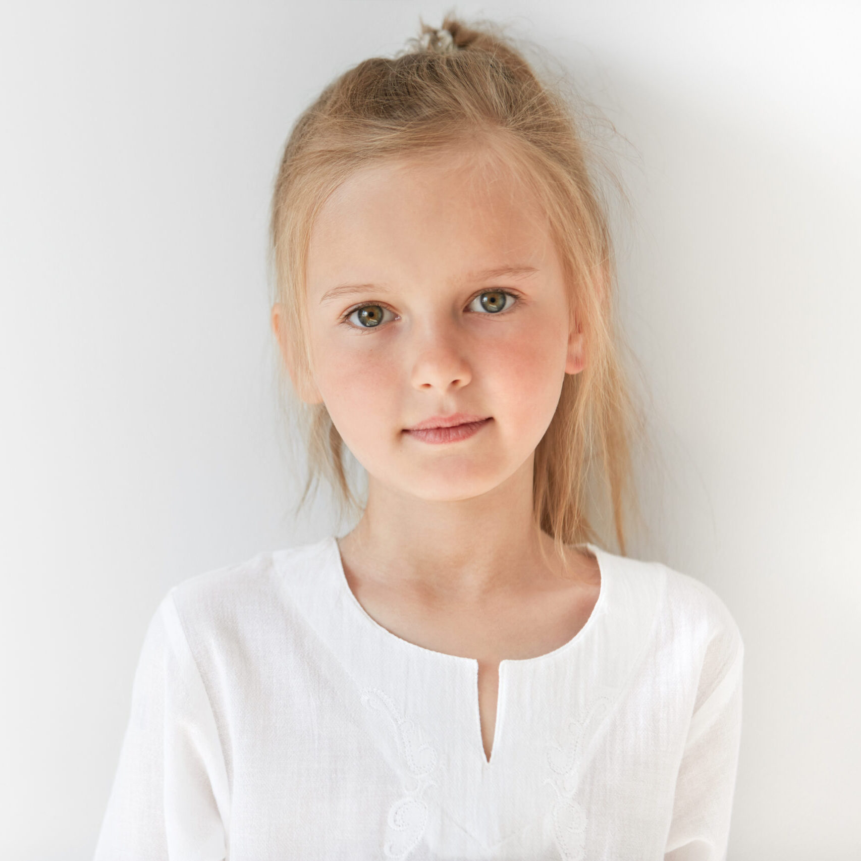 Blond Caucasian female child with green eyes standing quietly indoors and looking straight forward in good and sunny day. White colors make little girl look like a little angel or innocent baby.